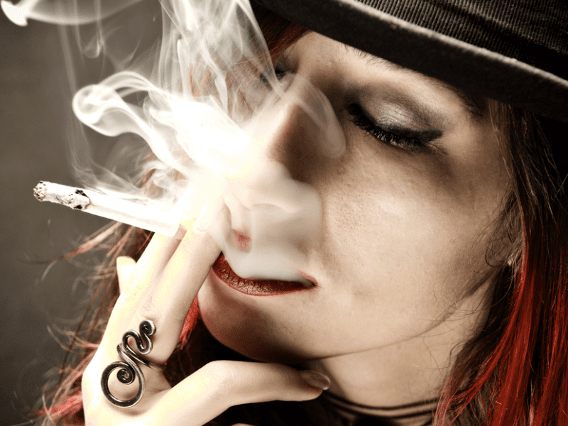 A woman wearing a stylish hat, casually holding a cigarette between her lips, exuding a confident demeanor.